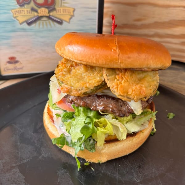 A tall Emerson Biggins burger with fried pickles, lettuce, tomato, and sauce on a dark plate.